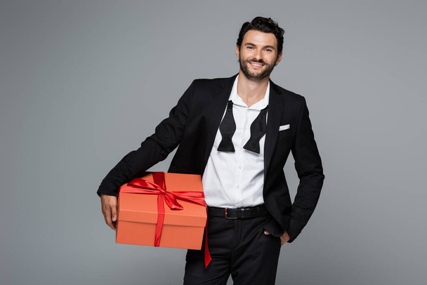 joyful man in suit holding red gift box and standing with hand in pocket isolated on grey  - Photo, Image