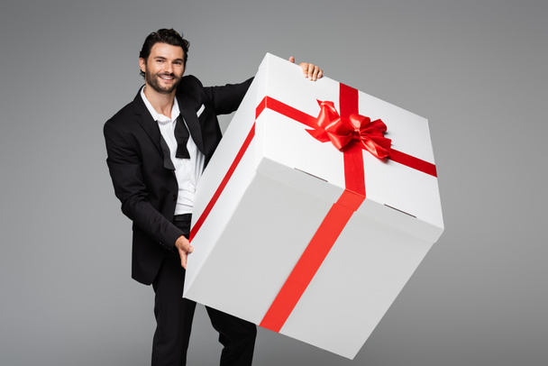 cheerful man in suit holding huge gift box isolated on grey  - Photo, Image