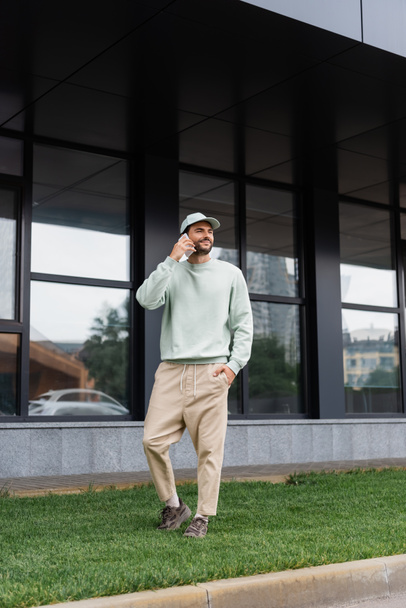 koko pituus tyytyväinen mies seisoo käsi taskussa ja puhuu matkapuhelin lähellä modernia rakennusta  - Valokuva, kuva
