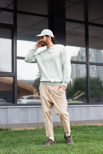 longitud completa del hombre sonriente de pie con la mano en el bolsillo y hablando en el teléfono celular cerca del edificio moderno  - Foto, imagen