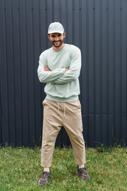 full length of smiling man in baseball cap posing with crossed arms near metallic fence  - Foto, immagini