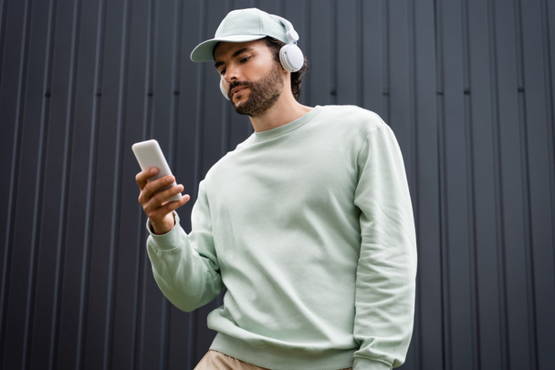 bearded man in cap listening music in wireless headphones and holding smartphone near metallic fence - Foto, afbeelding