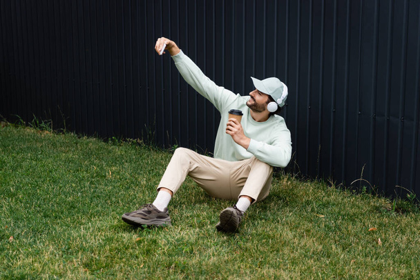 Kablosuz kulaklıklı mutlu adam yeşil çimlerde kağıt bardakla otururken selfie çekiyor.  - Fotoğraf, Görsel