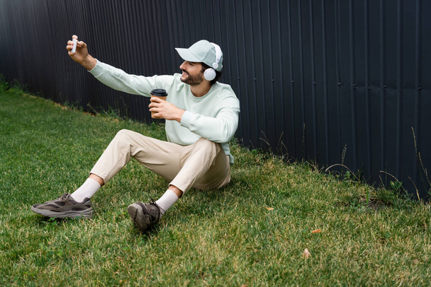 uomo positivo in cuffie wireless scattare selfie mentre seduto su erba verde con tazza di carta  - Foto, immagini