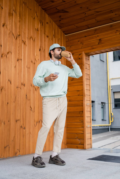 bärtiger Mann mit Mütze trinkt Coffee to go, während er sein Smartphone in der Nähe des Gebäudes hält  - Foto, Bild