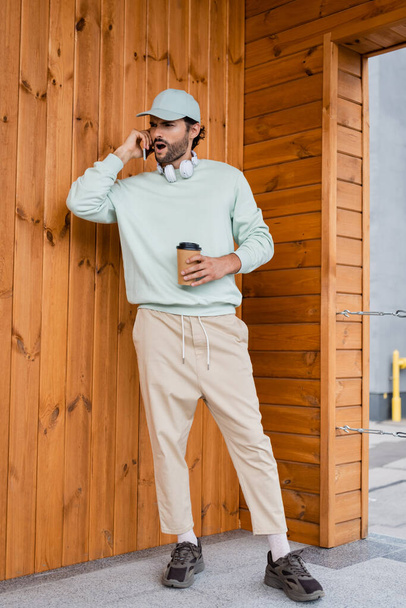 longitud completa del hombre sorprendido en la gorra de béisbol sosteniendo café para llevar mientras habla en el teléfono inteligente cerca del edificio  - Foto, imagen
