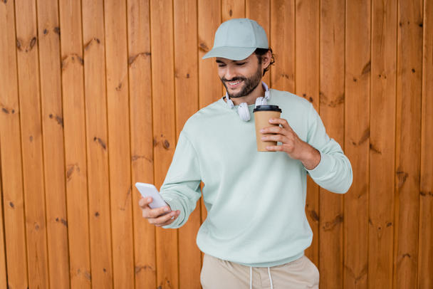 счастливый человек в кепке, держащий кофе, чтобы пойти, пользуясь смартфоном  - Фото, изображение