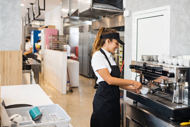 Beautiful and happy young female worker working in a bakery or fast food restaurant and using coffee machine. Positive people in small business concept. - 写真・画像