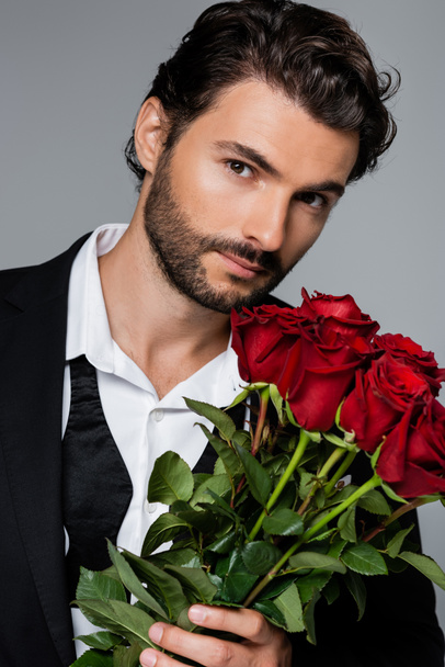 szakállas férfi öltönyben, vörös rózsákat tart a szürkén. - Fotó, kép