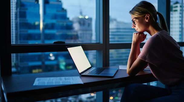 Side view of concentrated female manager sitting at table and looking at screen of laptop while doing work task in evening - 写真・画像