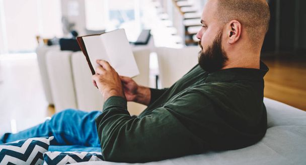 Side view of crop focused male in casual clothes and with beard sitting on sofa with diary and reading notes at home - Φωτογραφία, εικόνα