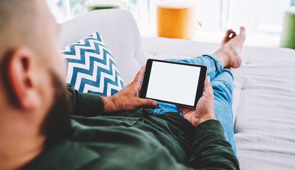 High angle of crop faceless bearded man in casual wear lying on sofa with crossed legs and watching movie on tablet while spending time alone on weekend at daytime - Fotó, kép