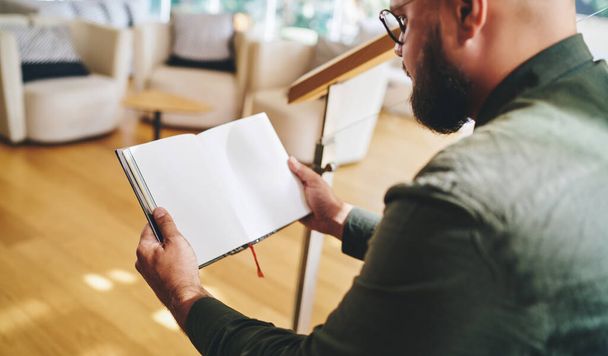 Side view of crop bearded man in casual wear and eyeglasses concentrating on reading book with blank pages while spending weekend in modern living room - Foto, imagen