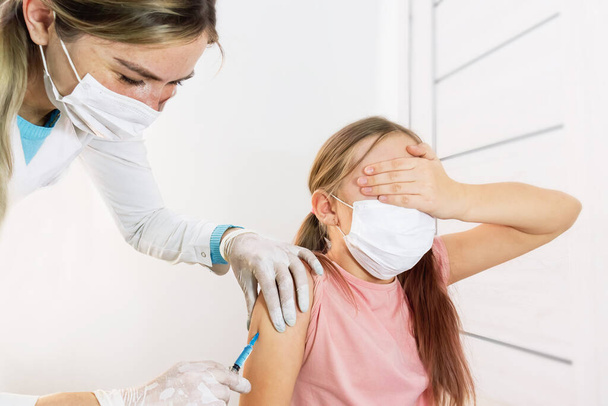 μια νοσοκόμα με ιατρική μάσκα και στολή κάνει έναν εμβολιασμό στον ώμο ενός κοριτσιού που έκλεισε τα μάτια της με το χέρι της από φόβο. Επιλεκτική εστία.Εμβολιασμός των παιδιών. Εμβολιασμοί και ενέσεις παιδιών στο ιατρείο. - Φωτογραφία, εικόνα