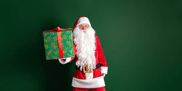 Emotionaler Schauspieler im Kostüm des Weihnachtsmannes hält Geschenkbox auf grünem Hintergrund in die Kamera - Foto, Bild