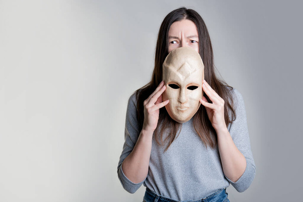 A young woman holds a mask in her hands, hiding her face half, a frightened look over the mask. - Valokuva, kuva