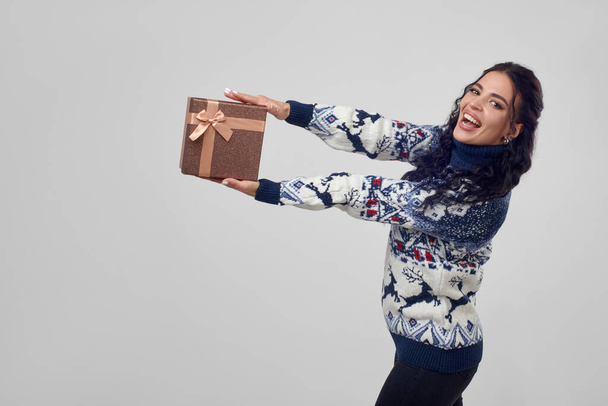 Pretty woman holds out a gift box on a white background, in a Christmas sweater. - Foto, afbeelding