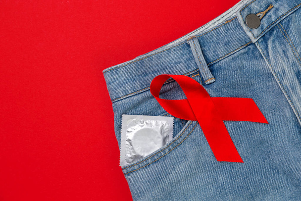 Jeans with a condom in the pocket and a red ribbon. Red background. Flat lay. Copy space. The concept of world AIDS day and safe sex. - Photo, image