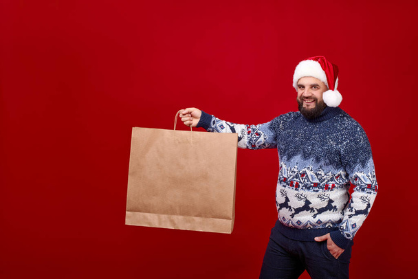 Мужчина держит большую сумочку на красном фоне в новогодней кепке.. - Фото, изображение