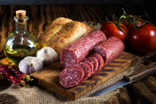 salami on a cutting board with slices. Garlic, peppers and tomatoes on a wooden table. - Fotó, kép