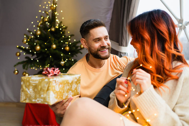 Молода пара сидить у затишному гламурі. Чоловік дарує жінці подарунок на Різдво. Різдвяна ялинка на фоні
. - Фото, зображення