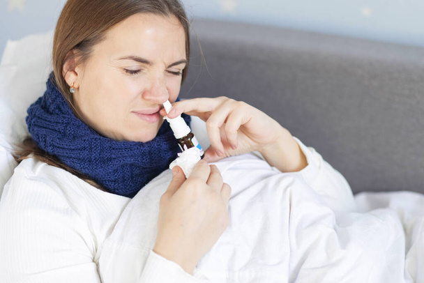 Een jonge vrouw met een zware verkoudheid ligt in bed onder de deken en gebruikt neusdruppels. Vrouw die neusspray gebruikt. - Foto, afbeelding