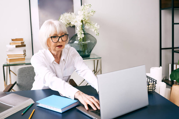 cansado senior hermosa mujer de pelo gris en blusa blanca trabajando en el ordenador portátil en la oficina. Trabajo, personas mayores, problemas, encontrar una solución, concepto de experiencia - Foto, Imagen
