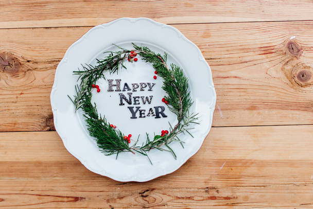 Happy New Year porcelain plate with Christmas wreath. Party decoration, flat lay, top view. Empty space. Restaurant menu concept. Isolated on wooden table background. Winter holidays background. - Foto, Imagem