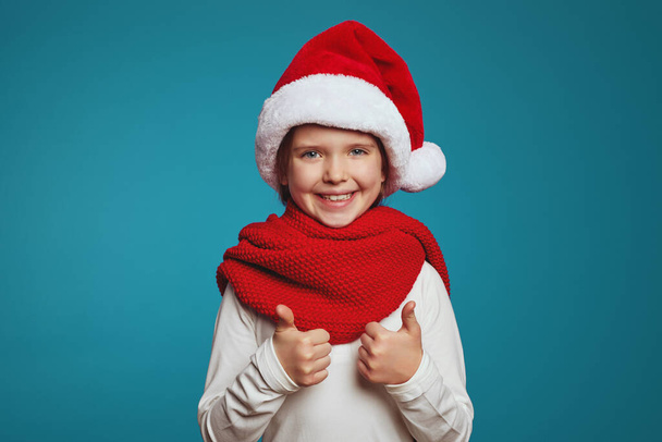 Cheerful little girl wearing christmas hat and red scarf, showing thumbs up - Foto, Bild