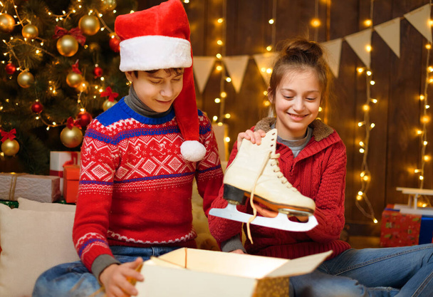 Portrait of a boy and girl in New Year decoration. They open a box, take out their skates and have fun. Holiday lights, gifts and a Christmas tree decorated with toys. - Photo, image