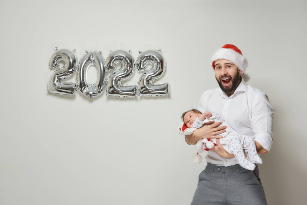 Egy piros bársony sapkás apa 2022-es alakban tartja a kislányát ezüst lufik közelében. Egy szakállas apa a kisgyerekével kinyitja a száját egy újévi partin.. - Fotó, kép