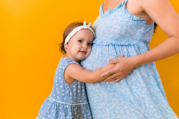 Motherhood, love, Childhood, hot summer - croped portrait pregnant mother unrecognizable woman blue dress little sibling girl sister hug touch mum big belly brother on yellow background copy space - Fotografie, Obrázek