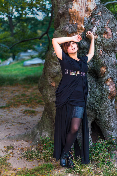 Μια Κινέζα, ντυμένη στα σκοτεινά, είναι κουρασμένη, δυστυχισμένη, στέκεται δίπλα σε ένα παλιό κορμό δέντρου το βράδυ. - Φωτογραφία, εικόνα