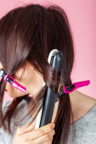 A beautiful young caucasian woman is using sectioning clips and heat hair iron wand for straightening or curling her hair. She is against pink background. DIY beauty concept with electircal tools. - Foto, Bild