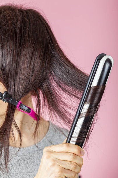 Egy gyönyörű, fiatal kaukázusi nő vágókapcsokat és hő hajvasaló pálcát használ a haja kiegyenesítésére vagy meghajlítására. Rózsaszín háttérrel rendelkezik. DIY szépség koncepció elektrotechnikai eszközökkel. - Fotó, kép