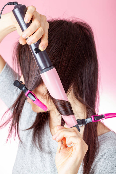 A beautiful young caucasian woman is using a conical electrical curler wand or curling iron to create curles in her red brown hair. DIY cosmetics concept against pink background. - Photo, Image