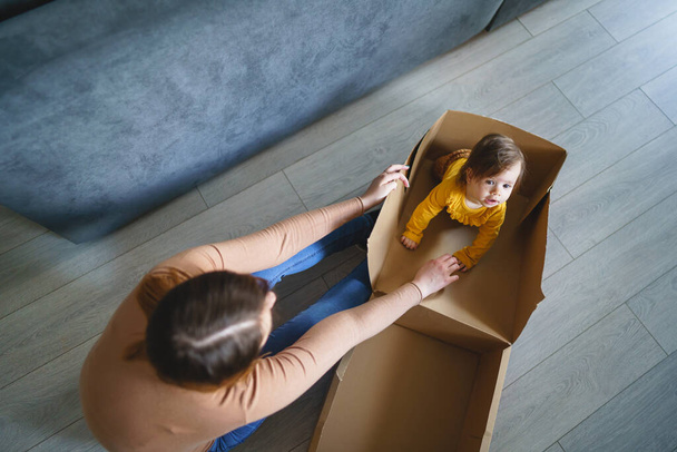 Una pequeña niña caucásica jugando por su madre en la caja de cartón en casa sentada en la caja de personas reales de la familia y creciendo concepto de copia espacio vista superior - Foto, Imagen