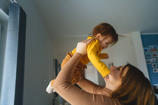 One small caucasian baby girl happy in hands of her mother holding her at home in day high up having fun real people side view copy space family bonding leisure concept - 写真・画像