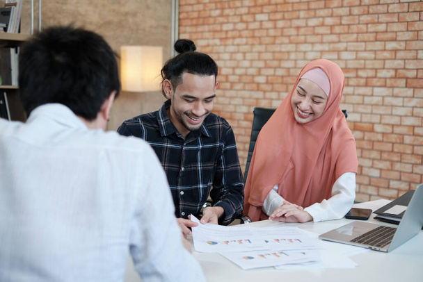 Dois jovens colegas de startup que são islâmicos falam sobre projetos financeiros com um cliente com um sorriso, sucesso de apresentação com um gráfico de negócios na mesa de um pequeno local de trabalho de escritório. - Foto, Imagem