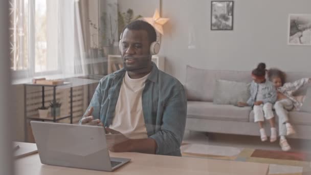 Medium slow-shot di uomo afroamericano in cuffia in videoconferenza su laptop da casa con due figlie giocose che si divertono in background - Filmati, video