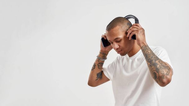Focused mixed race tattooed young man wearing headphones, listening to hip hop music while posing isolated over white background - 写真・画像
