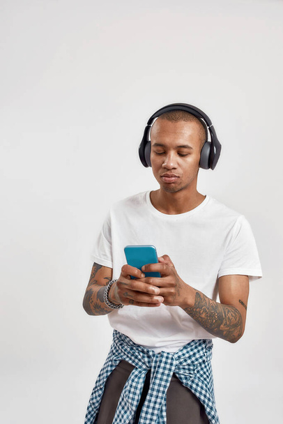 Portrait of busy mixed race young man wearing headphones sending and getting messages, using smartphone, standing isolated over light background - Valokuva, kuva