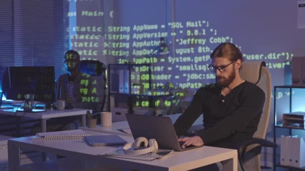 Střední záběr dvou různých mužských programátorů sedících na pracovišti v tmavé kanceláři s běžícími programovacími kódy promítajícími do interiéru kanceláře - Záběry, video