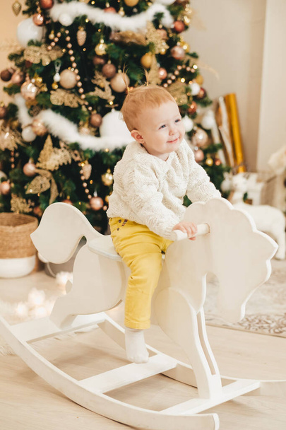 A blonde girl in a sweater rides a wooden horse at home against the background of the Christmas tree. Cute little girl playing at home on the background of garlands. - Photo, Image