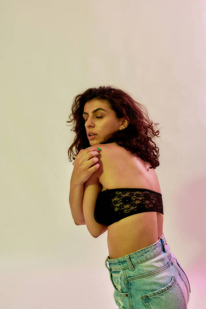 Seductive female model with curly dark hair in black underwear top posing isolated over neon light background - 写真・画像