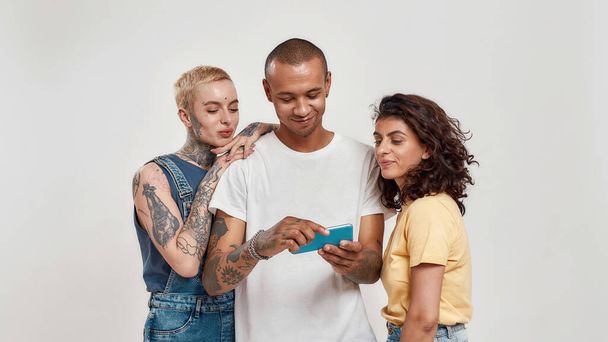 Three young diverse people in casual wear using smartphone, watching video, standing together isolated over light background - 写真・画像