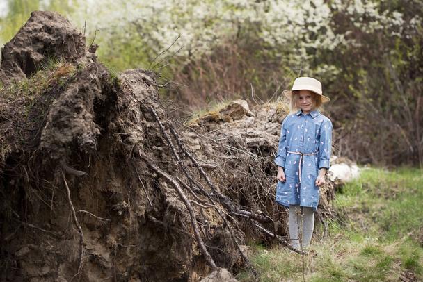 Portrait of little caucasian girl standing next to a fallen tree with upturned roots in the forest and looks at the camera - Foto, Bild