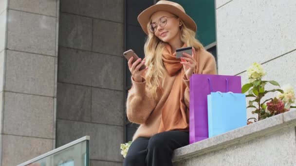 Millennial chica haciendo compras en línea usando teléfono y tarjeta de crédito asiente cabeza afirmativa hembra custimer felizmente bailando caucásico mujer sonriendo recibiendo cashback sentado al aire libre con coloridas bolsas - Metraje, vídeo