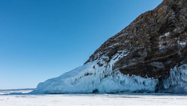 Een granieten rots, verstoken van vegetatie, rijst uit boven een bevroren meer. Er zijn scheuren op de pistes. Er is een dikke laag ijspegels aan de basis. Heldere blauwe lucht. Baikal. - Foto, afbeelding
