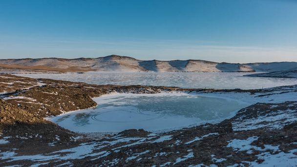 Een klein bevroren meer in de vorm van een hart wordt van de grote gescheiden door een strook land. Sneeuw op de oevers. Een bergketen tegen een blauwe lucht. Baikal. - Foto, afbeelding